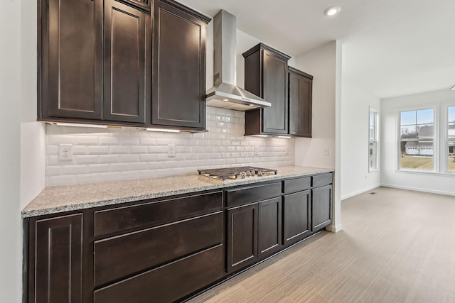 kitchen with stainless steel gas cooktop, dark brown cabinets, light stone countertops, decorative backsplash, and wall chimney range hood