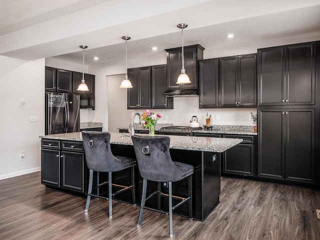 kitchen with pendant lighting, dark hardwood / wood-style flooring, light stone counters, a kitchen island with sink, and black fridge