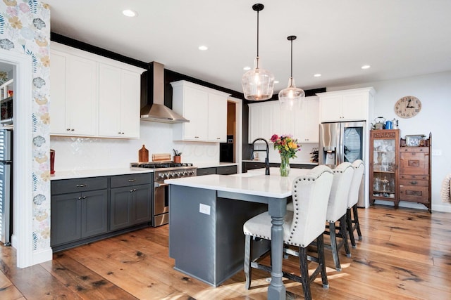 kitchen featuring wall chimney exhaust hood, white cabinetry, decorative light fixtures, a center island with sink, and appliances with stainless steel finishes