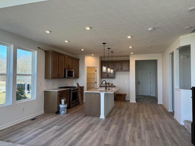 kitchen featuring an island with sink, light stone counters, light wood-style floors, appliances with stainless steel finishes, and baseboards