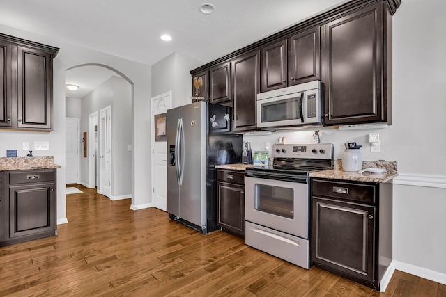 kitchen featuring stainless steel appliances, dark brown cabinets, dark wood-type flooring, and light stone counters