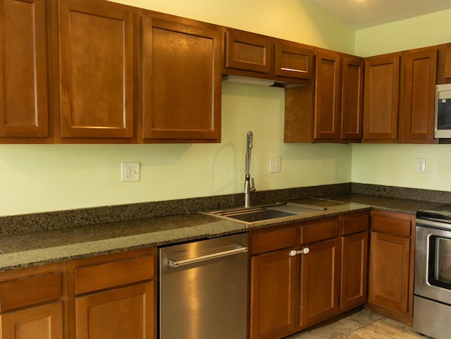 kitchen with dark stone counters, stainless steel appliances, brown cabinetry, and a sink