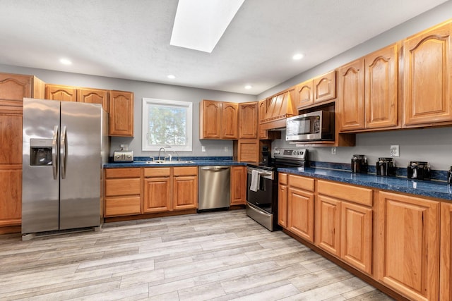 kitchen with a skylight, dark countertops, appliances with stainless steel finishes, light wood-style floors, and a sink