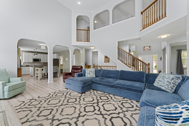 living area featuring arched walkways, light wood-style flooring, a towering ceiling, and stairs