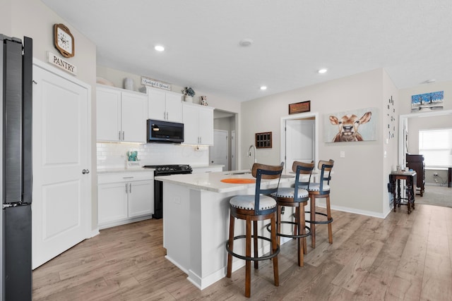 kitchen featuring white cabinetry, an island with sink, backsplash, black appliances, and light wood-type flooring