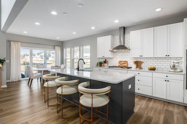 kitchen featuring a breakfast bar area, white cabinets, a kitchen island with sink, stainless steel gas cooktop, and wall chimney range hood