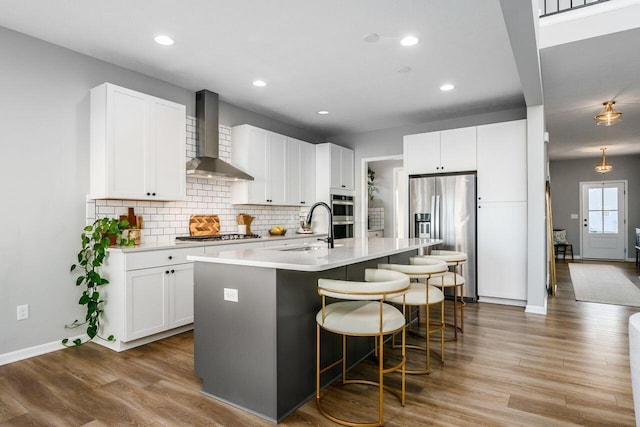 kitchen featuring white cabinetry, stainless steel appliances, a center island with sink, and wall chimney range hood