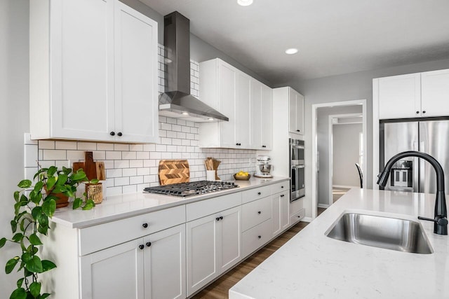 kitchen with appliances with stainless steel finishes, sink, white cabinets, light stone counters, and wall chimney range hood