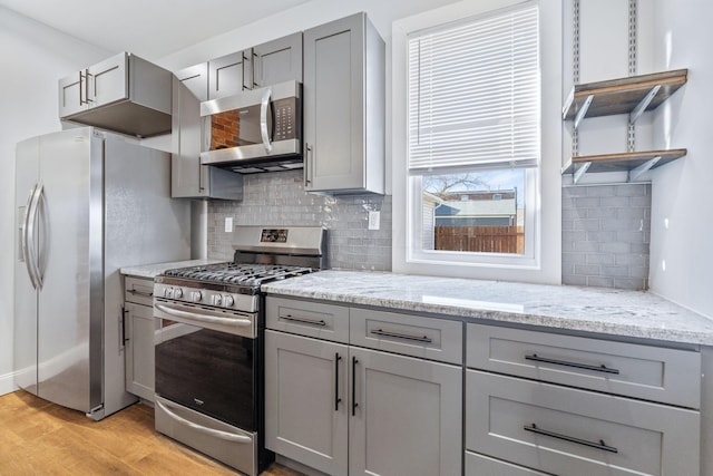 kitchen featuring appliances with stainless steel finishes, tasteful backsplash, gray cabinetry, light stone countertops, and light hardwood / wood-style flooring