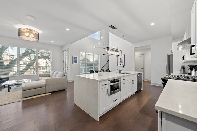 kitchen with stainless steel appliances, light stone counters, an island with sink, white cabinets, and decorative light fixtures