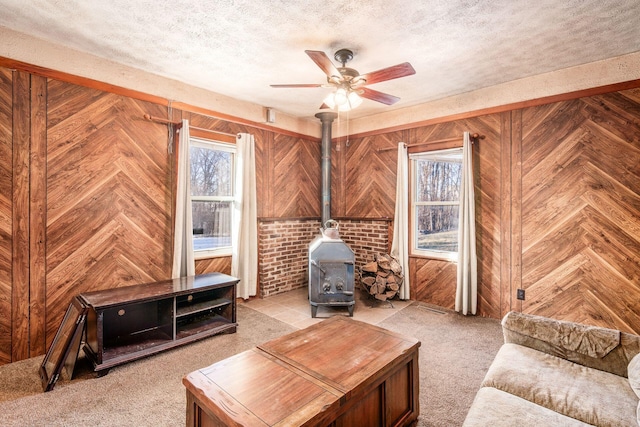 carpeted living room with ceiling fan, a wood stove, a textured ceiling, and wood walls