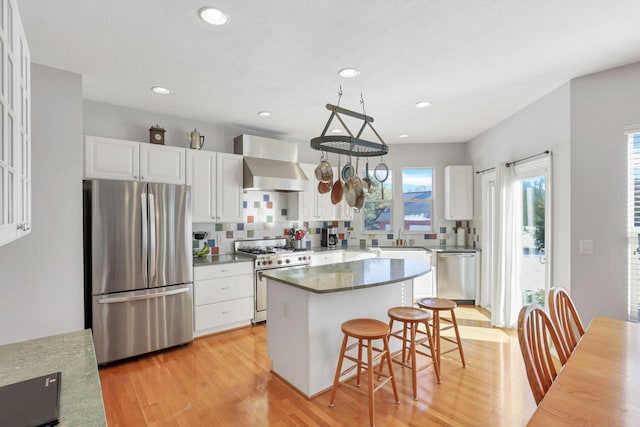 kitchen with a kitchen island, appliances with stainless steel finishes, white cabinetry, a kitchen bar, and wall chimney range hood