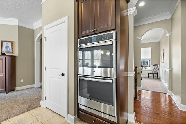 kitchen with crown molding, stainless steel double oven, light colored carpet, and dark brown cabinets