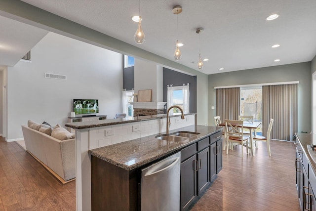 kitchen with decorative light fixtures, dishwasher, sink, dark stone countertops, and a kitchen island with sink