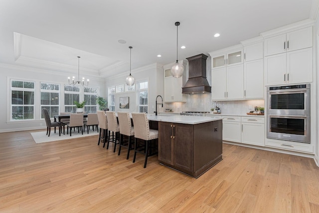 kitchen featuring decorative light fixtures, appliances with stainless steel finishes, custom range hood, a kitchen island with sink, and white cabinets