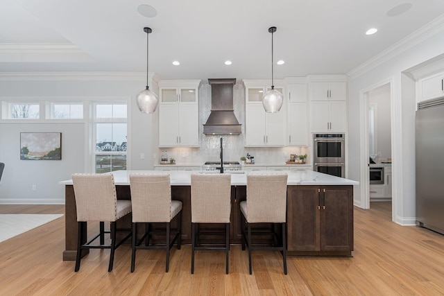 kitchen featuring custom exhaust hood, white cabinetry, appliances with stainless steel finishes, pendant lighting, and a kitchen island with sink