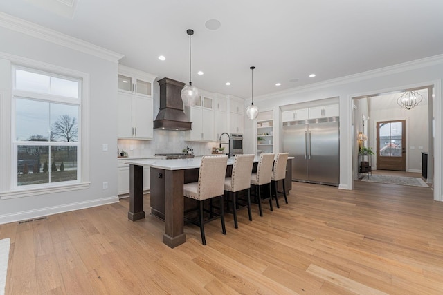 kitchen with appliances with stainless steel finishes, a kitchen island with sink, hanging light fixtures, white cabinetry, and custom range hood