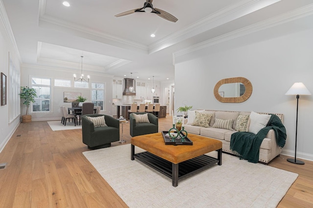 living room with ornamental molding, a raised ceiling, ceiling fan with notable chandelier, and light wood-type flooring