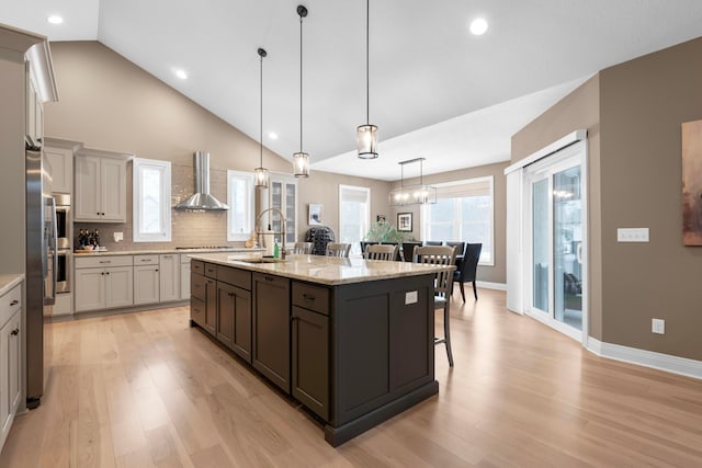 kitchen with decorative light fixtures, sink, a kitchen island with sink, light stone countertops, and wall chimney range hood