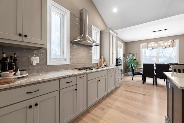 kitchen featuring wall chimney range hood, hanging light fixtures, light stone countertops, stainless steel gas cooktop, and vaulted ceiling