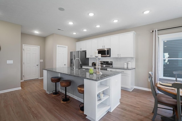 kitchen featuring appliances with stainless steel finishes, an island with sink, hardwood / wood-style flooring, dark stone counters, and white cabinets