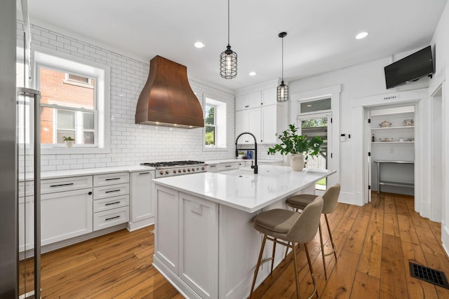 kitchen with premium range hood, white cabinetry, hanging light fixtures, light hardwood / wood-style flooring, and a kitchen island with sink