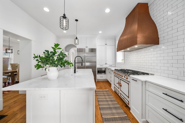 kitchen featuring premium range hood, appliances with stainless steel finishes, white cabinetry, hanging light fixtures, and a kitchen island with sink