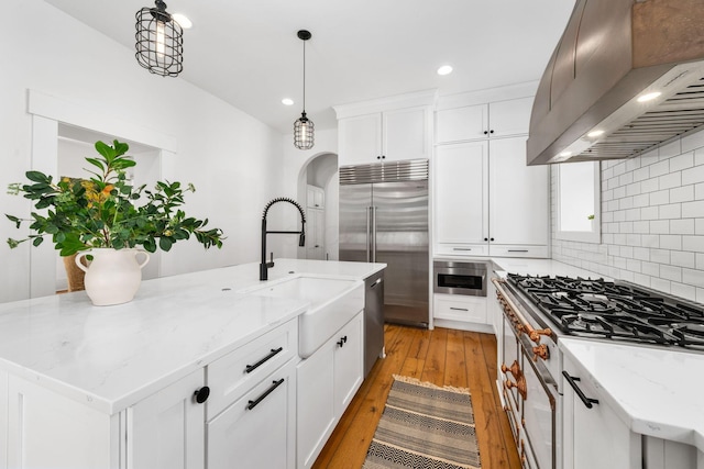 kitchen with a kitchen island with sink, premium appliances, white cabinets, decorative light fixtures, and exhaust hood