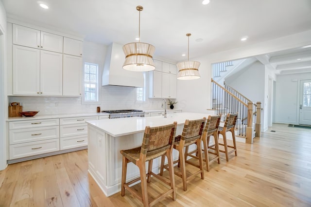 kitchen with premium range hood, an island with sink, pendant lighting, light hardwood / wood-style floors, and white cabinets