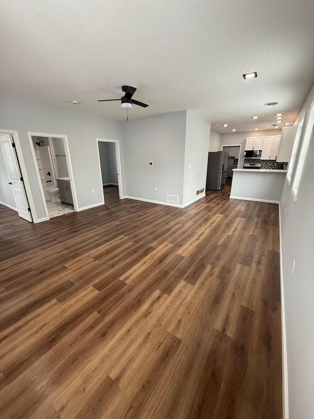 unfurnished living room with a textured ceiling, ceiling fan, visible vents, baseboards, and dark wood finished floors