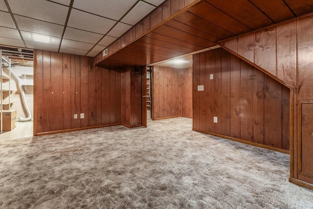 basement featuring light carpet, a drop ceiling, and wood walls