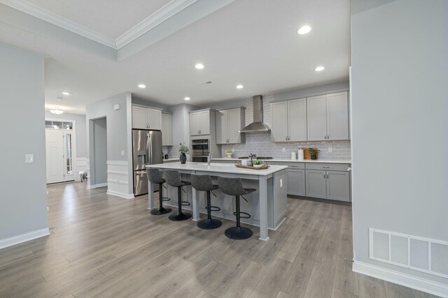 kitchen with wall chimney range hood, a breakfast bar, a kitchen island with sink, gray cabinetry, and stainless steel refrigerator with ice dispenser