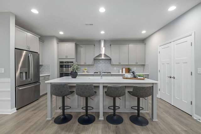 kitchen with a breakfast bar area, gray cabinetry, a center island with sink, stainless steel appliances, and wall chimney range hood