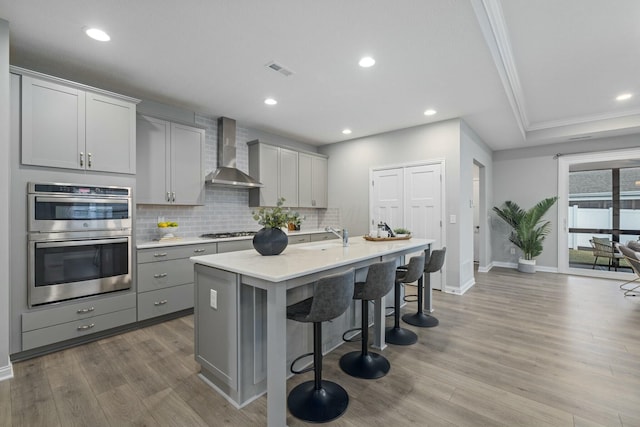 kitchen featuring wall chimney exhaust hood, a kitchen breakfast bar, gray cabinets, an island with sink, and stainless steel appliances