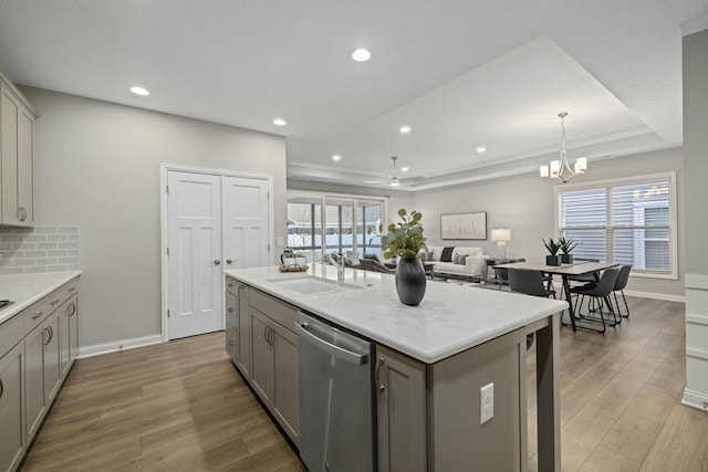 kitchen with gray cabinets, an island with sink, sink, stainless steel dishwasher, and a tray ceiling