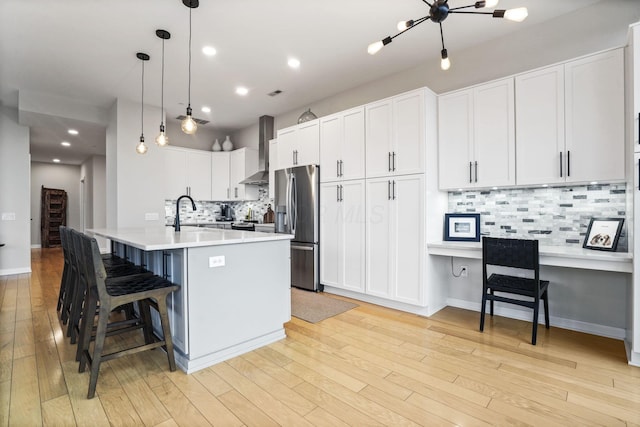 kitchen with a kitchen island with sink, white cabinets, stainless steel fridge with ice dispenser, decorative light fixtures, and wall chimney exhaust hood