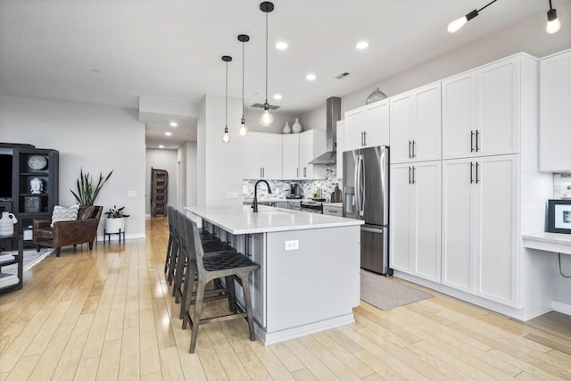 kitchen with appliances with stainless steel finishes, pendant lighting, wall chimney range hood, a kitchen island with sink, and white cabinets