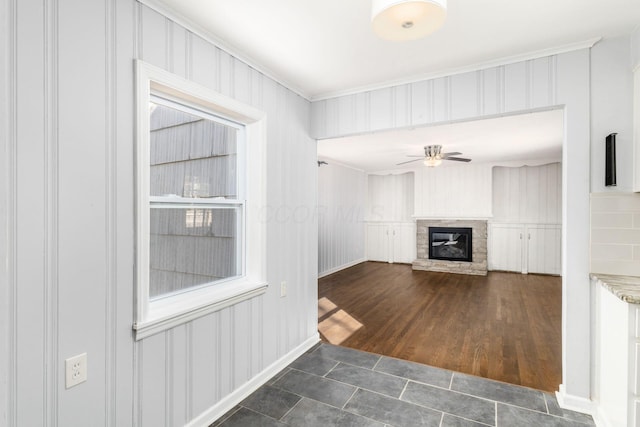 unfurnished living room with dark wood-type flooring, a stone fireplace, ceiling fan, and crown molding