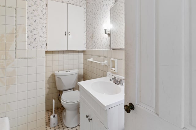 bathroom with wallpapered walls, a wainscoted wall, toilet, vanity, and tile walls