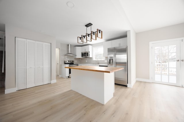 kitchen featuring butcher block countertops, wall chimney range hood, stainless steel appliances, a kitchen island, and decorative light fixtures