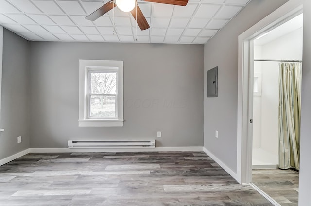 empty room featuring ceiling fan, baseboard heating, electric panel, wood-type flooring, and a drop ceiling