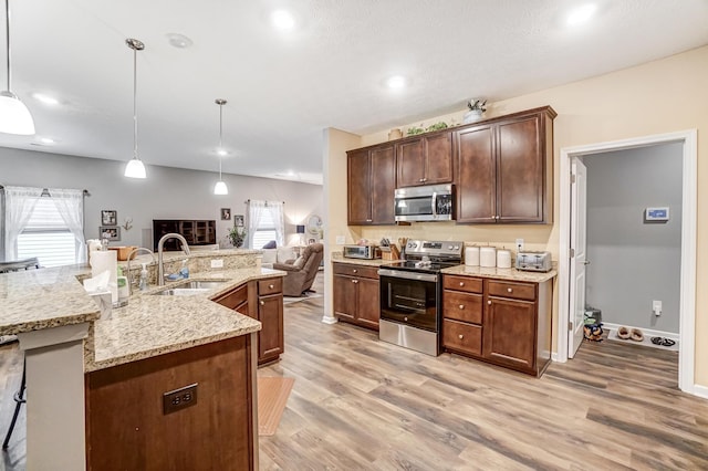 kitchen with sink, a kitchen island with sink, stainless steel appliances, decorative light fixtures, and light wood-type flooring