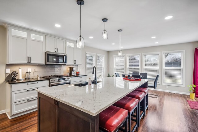 kitchen with appliances with stainless steel finishes, an island with sink, sink, white cabinets, and hanging light fixtures