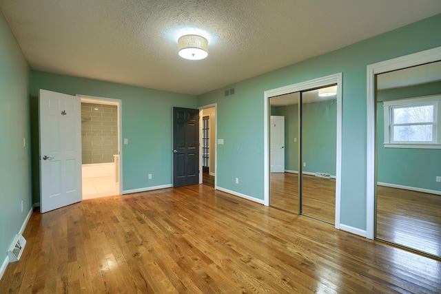 unfurnished bedroom featuring two closets, light hardwood / wood-style flooring, ensuite bath, and a textured ceiling