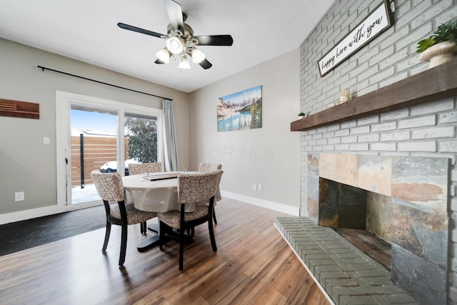 dining area featuring dark wood-style floors, a brick fireplace, baseboards, and a ceiling fan