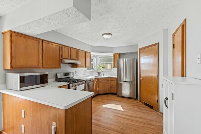 kitchen with light countertops, appliances with stainless steel finishes, brown cabinetry, a peninsula, and under cabinet range hood