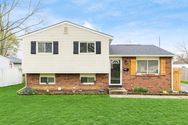 tri-level home featuring brick siding, fence, a front lawn, and roof with shingles