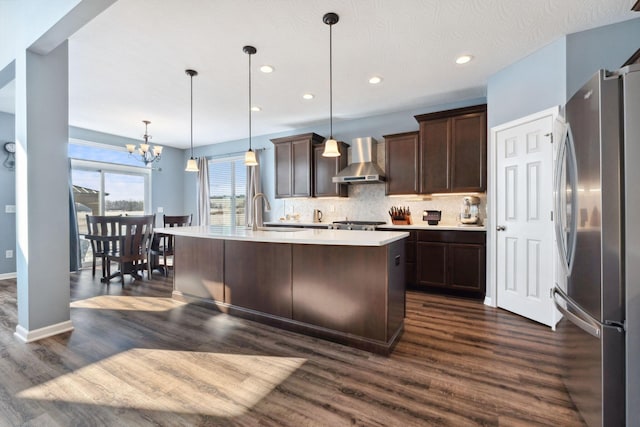 kitchen featuring light countertops, wall chimney range hood, dark brown cabinets, freestanding refrigerator, and decorative backsplash