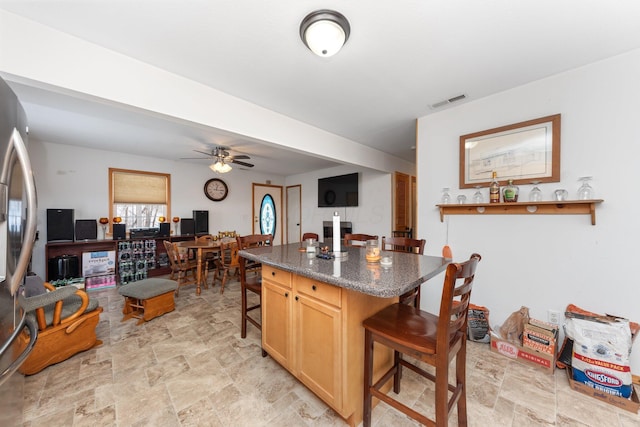 kitchen featuring stainless steel refrigerator, a kitchen breakfast bar, dark stone counters, a center island, and ceiling fan