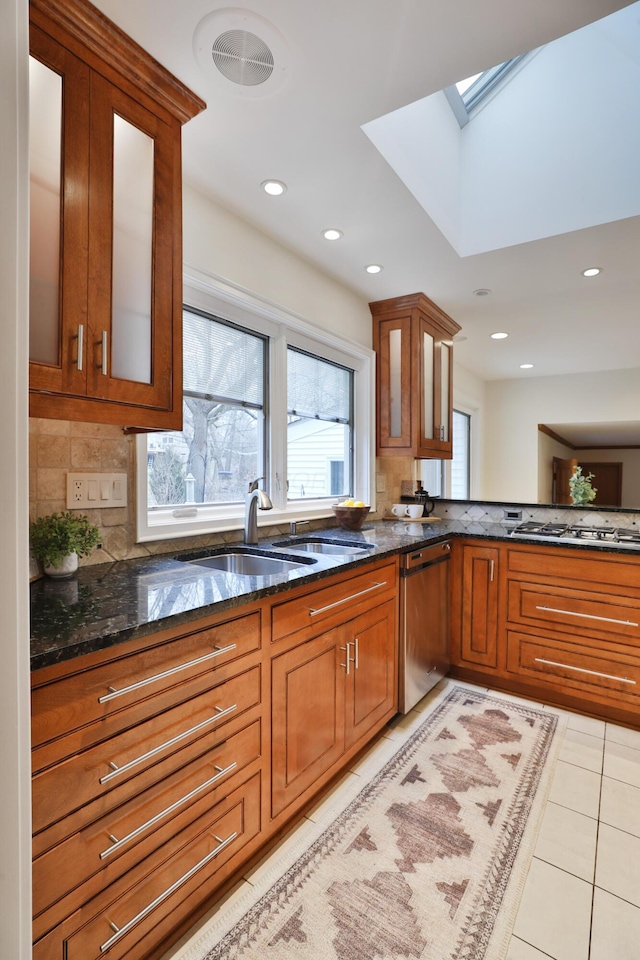 kitchen with a skylight, light tile patterned floors, stainless steel appliances, brown cabinetry, and dark stone countertops